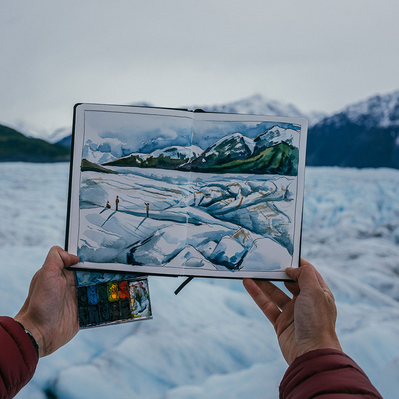 An artist holding a notebook in front of snowy mountains.